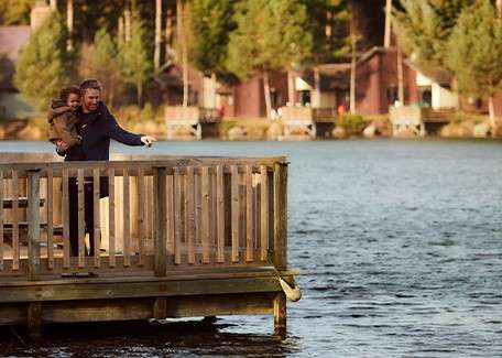 Adult holding a small child points toward the water, standing on a wooden dock. Across the lake, rustic cabins sit among tall evergreen trees in warm evening light.