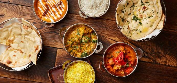 Assorted Indian dishes sit arranged on a wooden table: metal bowls of curries, yellow rice, and white rice, alongside naan breads, garnished with herbs and drizzled yogurt.