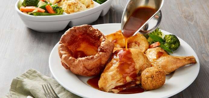 Gravy pours onto roast chicken on a plate, accompanied by Yorkshire pudding, roast potatoes, stuffing ball, broccoli and carrots; a side bowl of vegetables and cutlery rest on the table.