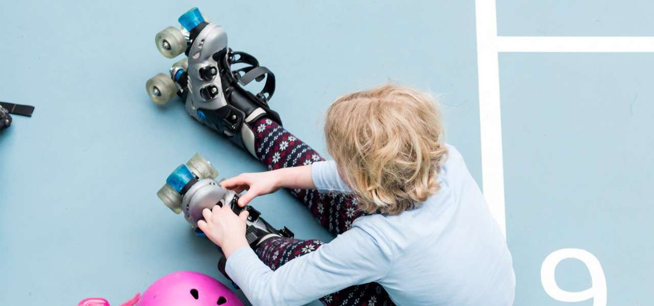 Child tightens gray quad roller skates while seated, near a pink helmet, on a light-blue court marked with white lines and the number 9.