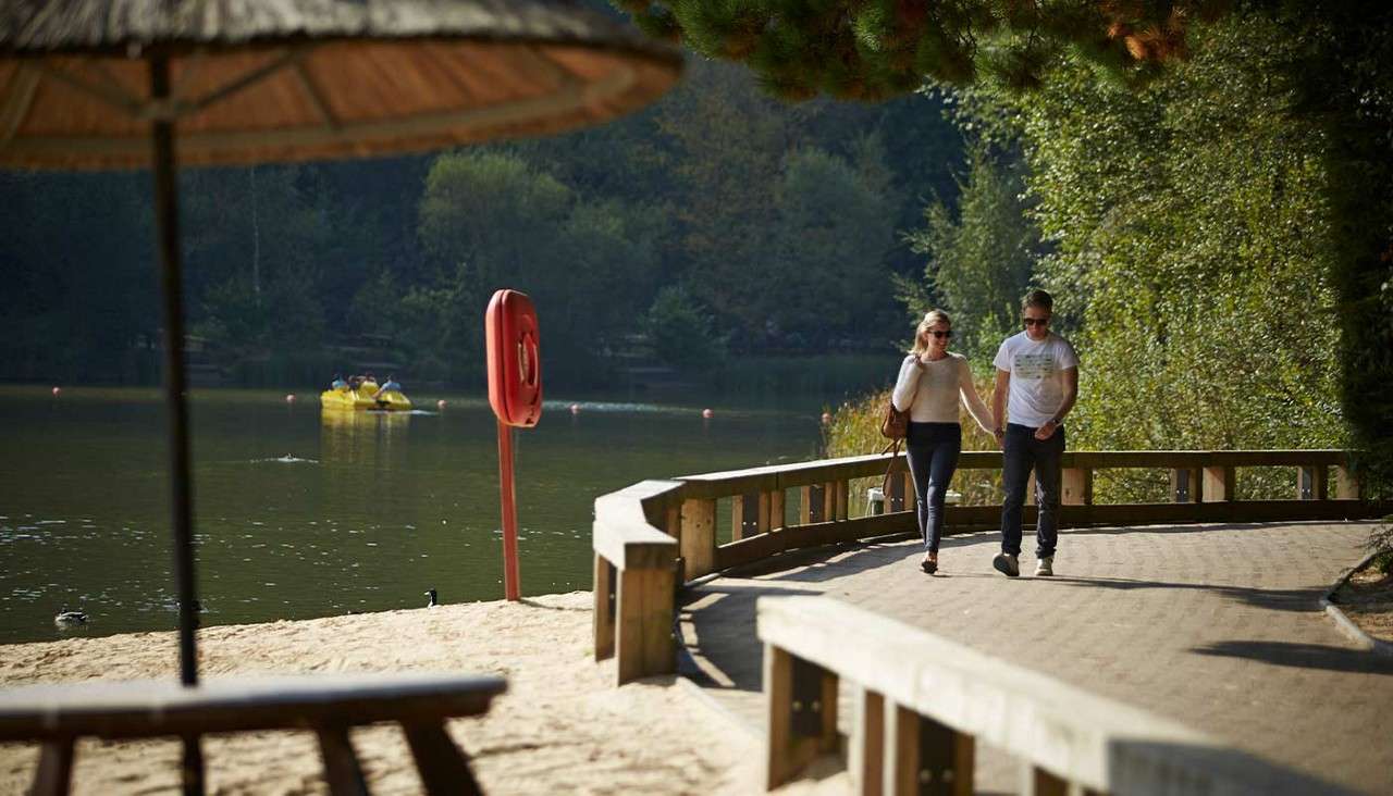 Couple walks hand-in-hand along a curved lakeside boardwalk, passing a red lifebuoy. In the background, people pedal a yellow boat on calm water beside a sandy beach and dense trees.