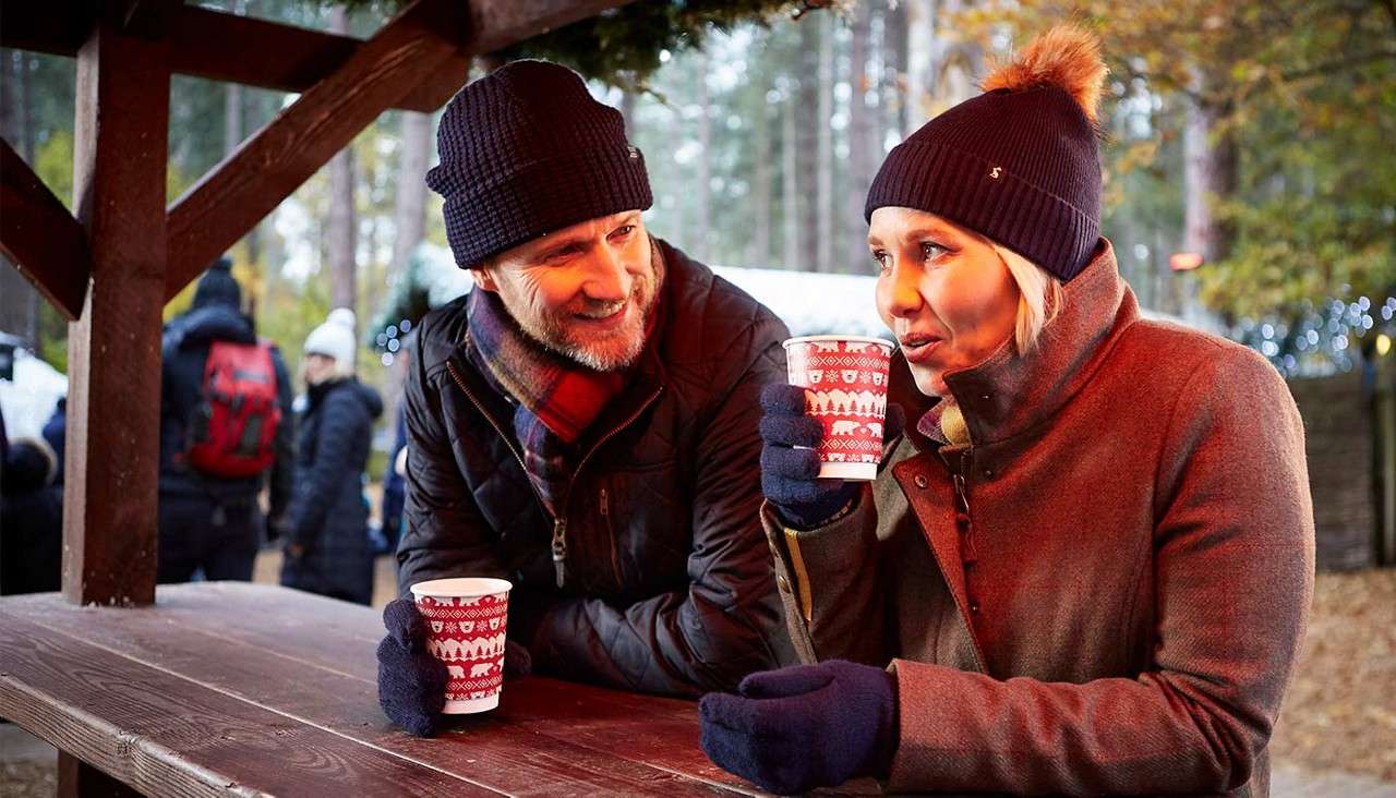 Two bundled adults drink hot beverages and chat, leaning on a wooden picnic table under a shelter, while people in coats pass through a wooded park with twinkling festive lights.