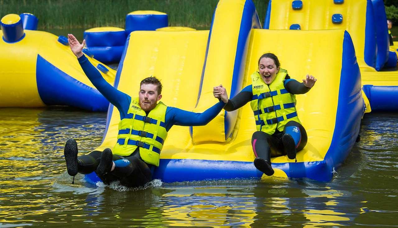 Two adults slide while holding hands, splashing into water from a yellow-blue inflatable water-park slide, wearing wetsuits and bright life vests.

Text: JOBE; L7; LZ.