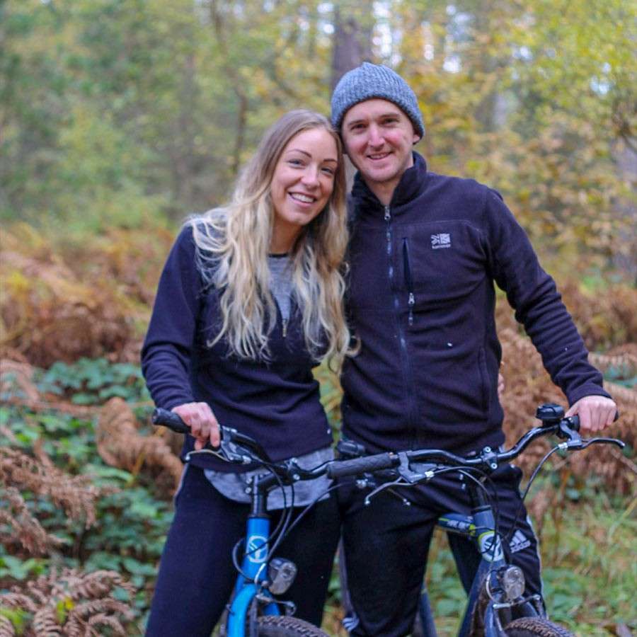 Two cyclists pose with mountain bikes, smiling and holding handlebars, in a wooded area. Fallen ferns and autumn foliage surround them on a forest trail.