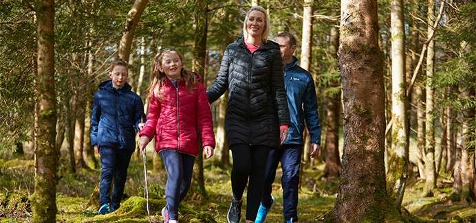 Four people wearing jackets walk and smile along a mossy forest trail, sunlight filtering through tall trees.