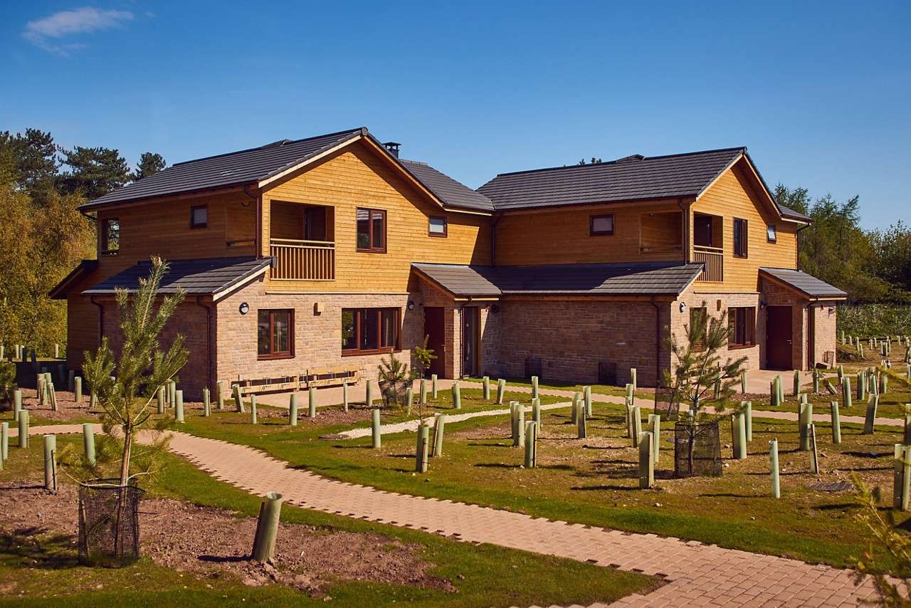 Two-story wooden-and-brick lodge sits amid landscaped grounds, with small balconies and dark-tiled roofs. Winding brick paths pass newly planted saplings in protective tubes, bordered by trees beneath clear blue sky.
