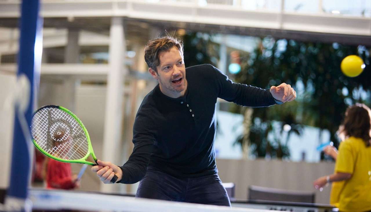 Man swings a green tennis racket at a yellow ball over a low net, leaning forward and focused, inside a spacious indoor atrium with blurred people, tables, and plants.