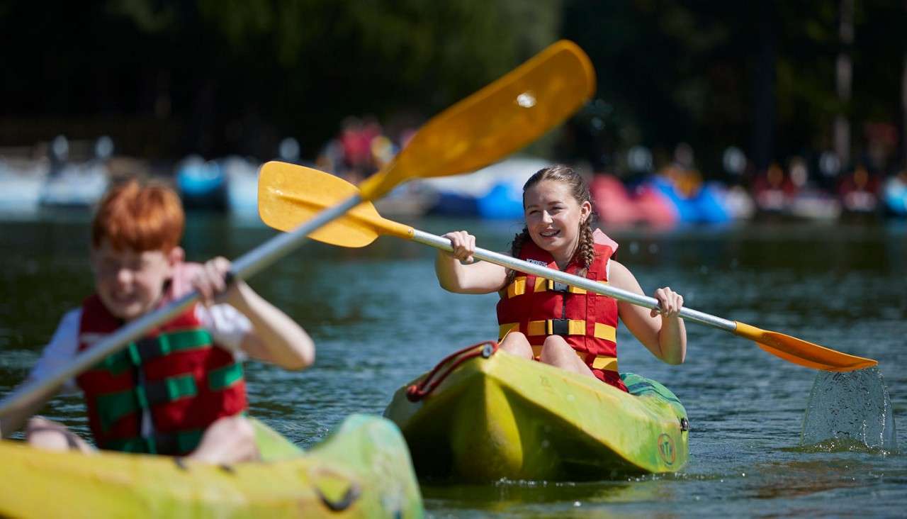 Two children paddle green kayaks, wearing red life jackets and using yellow oars, creating splashes on a calm lake, with blurred boats and trees in the sunny background.