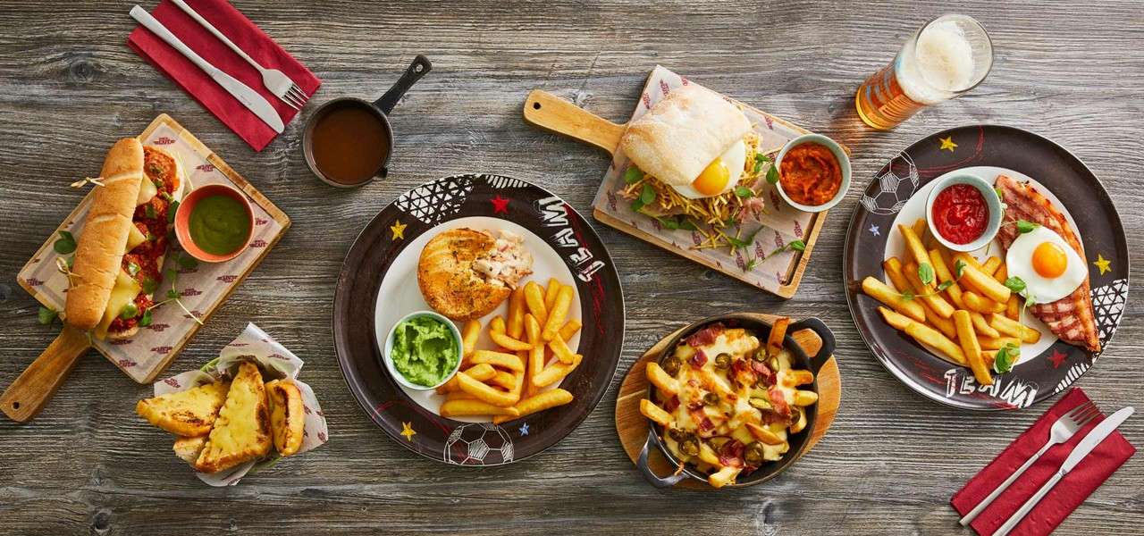 Assorted pub-style meals sit arranged on a wooden table: fries with dips, pies, eggs on toast, cheesy fries, and a meatball sub, with drinks and utensils nearby. Text: TEAM, TEAM.