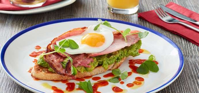 Toast topped with mashed avocado, bacon, and a sunny-side-up egg rests on a white plate, garnished with microgreens and hot sauce, on a wooden table beside orange juice and cutlery.