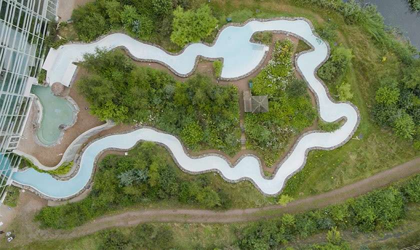 A serpentine lazy river winds around dense landscaped gardens, encircling trees and a gazebo; the water channel skirts a glass building and footpath within a park-like setting.