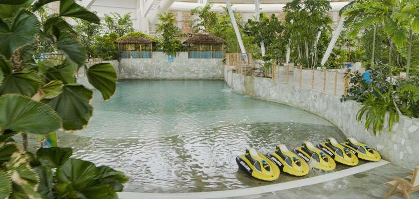 Yellow pedal boats rest side-by-side on a shallow edge, facing a calm indoor lagoon, surrounded by tropical plants, stone walls, and thatched huts beneath a bright, airy roof.