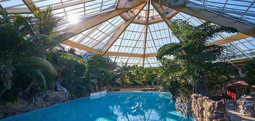 Indoor swimming pool reflects sunlight beneath a large glass-domed roof. Surrounding palm trees and rocks frame the water, with seating and pathways around a tropical-themed leisure complex.