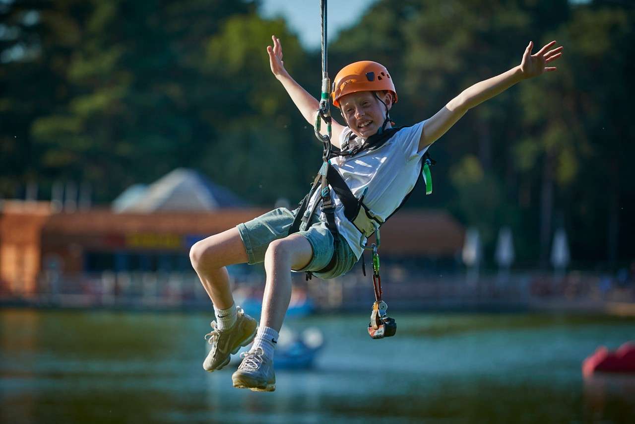 Two helmeted riders glide side-by-side on zip lines, legs splayed and hands gripping tethers, suspended by harnesses above a wooded landscape on an overcast day.