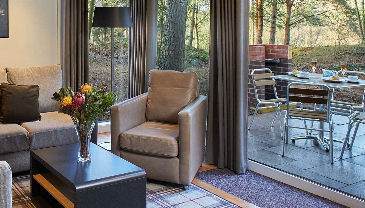 Vase of flowers rests on a coffee table, beside a leather armchair and sofa, while floor-to-ceiling windows reveal a patio dining set and brick grill amid trees.