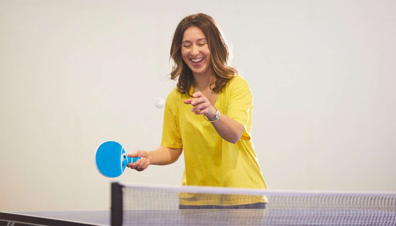 A woman swings a blue paddle to hit a ping-pong ball, smiling during play, beside a table tennis net in a bright indoor room with plain walls.