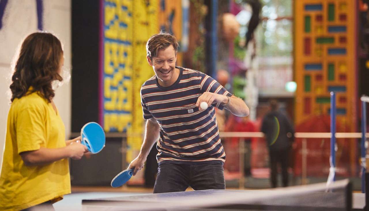 Man with striped shirt serves a ping-pong ball, smiling, while opponent in yellow waits with paddle, indoors at a colorful recreation center with climbing walls and blurred people.
