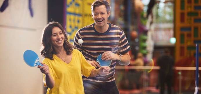Two adults with blue paddles volley a ping-pong ball, smiling and moving forward, in a brightly lit indoor recreation center with colorful play structures and blurred background activity.