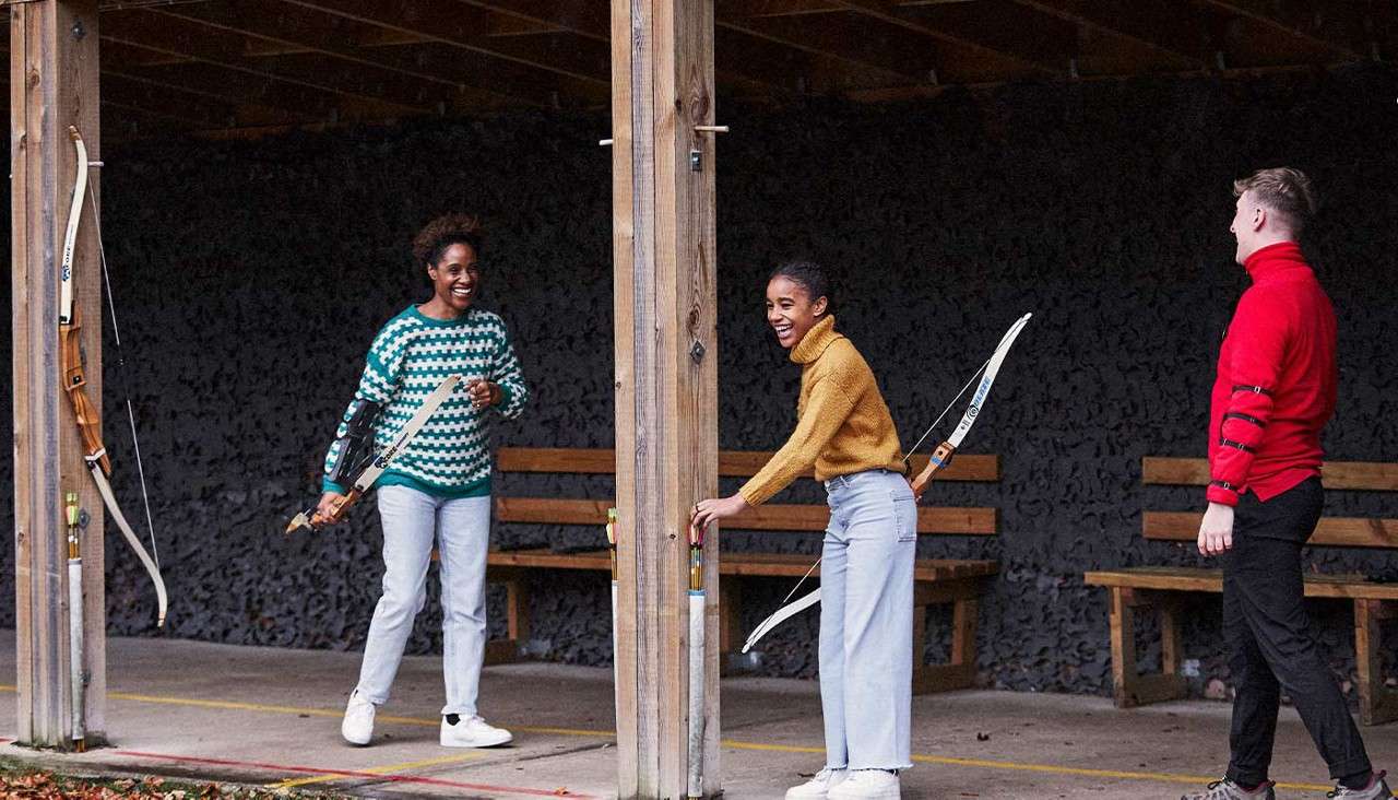 Three archers chat while holding recurve bows. They stand under a wooden, covered archery range with benches, hanging bows, yellow lane lines, and scattered leaves on the ground.
