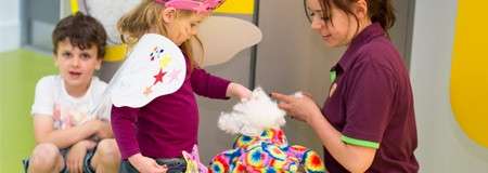 Child dresses a stuffed animal while an adult assists; another child watches nearby, sitting on the floor; scene in a bright, colorful indoor play or learning space.
