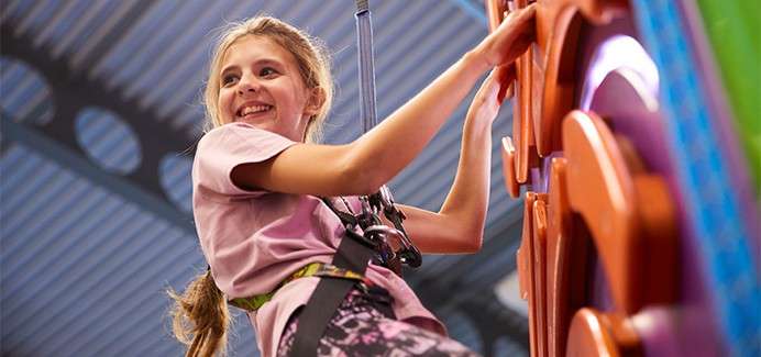 Child climber ascends a colorful indoor climbing wall, gripping molded holds while secured to a harness and auto-belay rope; smiling upward beneath a high, ribbed ceiling in a recreational facility.