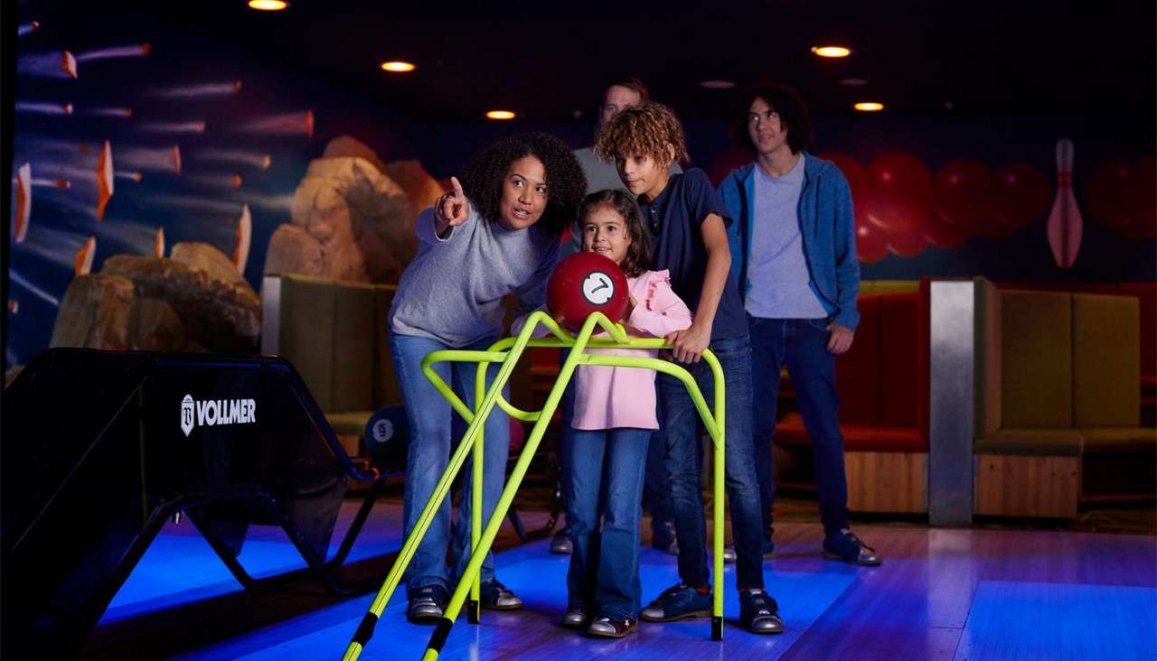 Child uses a bright-green bowling ramp to aim a red ball, guided by an adult, while others watch in a dim bowling alley near a ball return labeled T1 VOLLMER. Additional visible text: 9.
