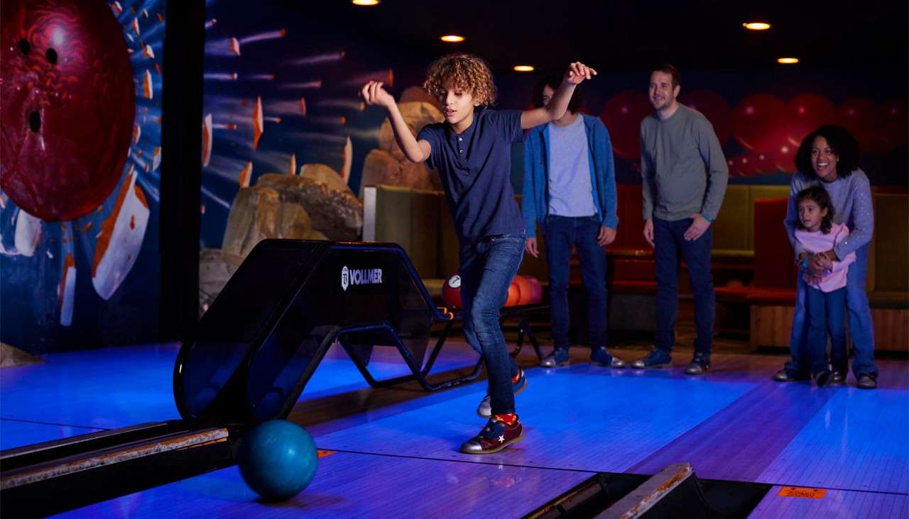 Child bowls; they release a blue ball down a lane while family watches nearby; indoor bowling alley with glowing lanes and a mural; ball return shows text: VOLLMER.