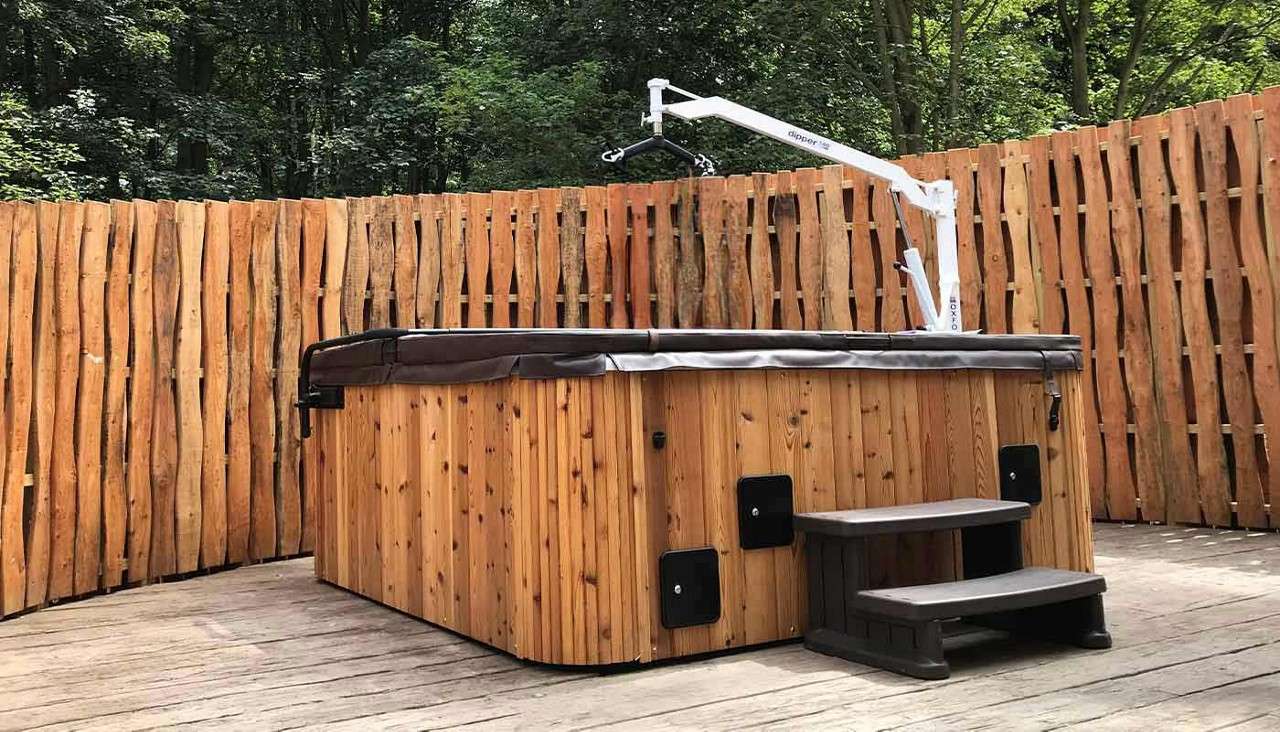 Wooden hot tub rests with cover closed, accessed by a white hoist labeled “dipper” and “OXFORD” and two plastic steps, within a curved rustic plank fence on a deck amid trees.