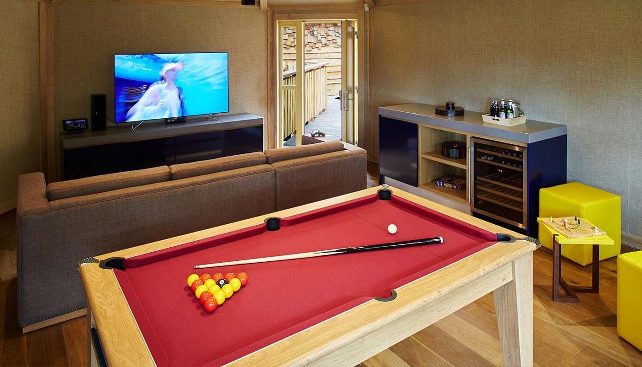 Pool table holds racked balls and cue, awaiting a break; surrounded by sofa facing TV, bar cabinet with drinks and wine fridge, yellow stools, and open doors leading to deck.