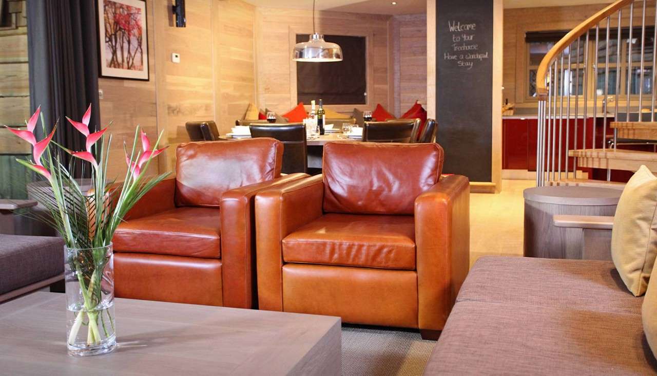 Two tan leather armchairs sit side by side, facing a coffee table with a flower vase, in an open-plan living-dining room. Chalkboard text: “Welcome to your Penthouse Have a wonderful Stay”.