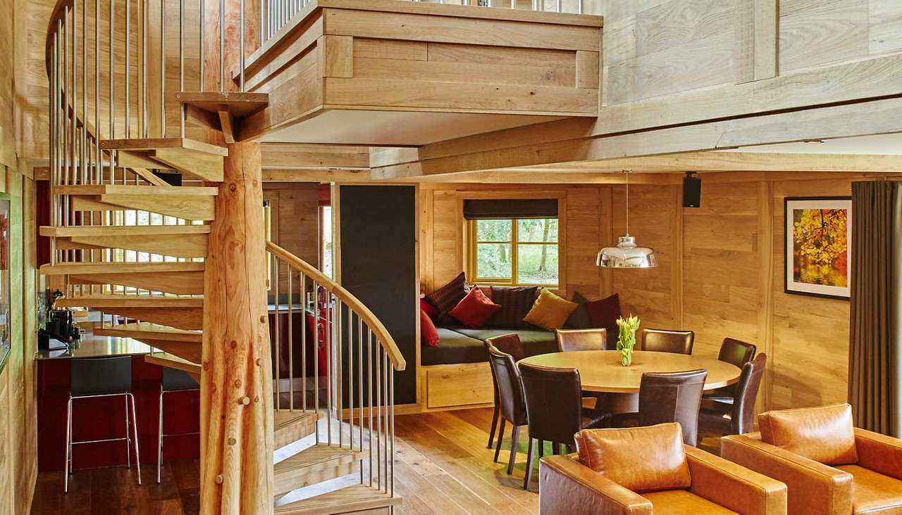 Spiral wooden staircase curves upward, anchored to a timber post; surrounding open-plan cabin shows leather armchairs, round dining table under pendant light, window seat with cushions, and adjacent kitchen bar.