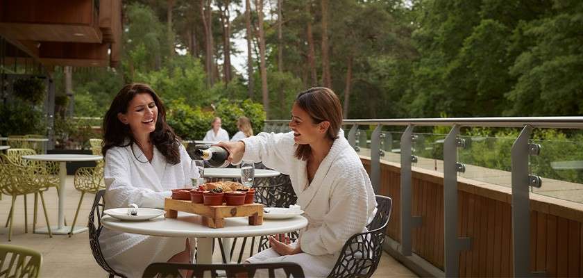 Two women in white spa robes sit at a patio table; one pours sparkling wine as they share snacks, relaxing on a terrace with chairs, glass railing, and forest backdrop.