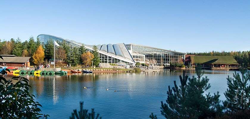 Glass-roofed leisure complex overlooks a calm lake, reflecting sunlight; pedal boats line the shore, ducks drift on the water, and forested buildings and evergreens surround under a clear blue sky.