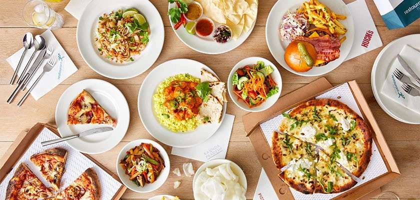 Assorted dishes rest arranged on a wooden table: two pizzas, pasta, curry over rice, salad, burger with fries, sauces, and drinks; nearby paper reads "HUGO'S" with cutlery and napkins.
