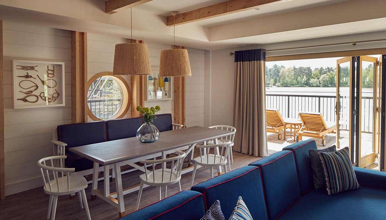 Dining table centers the room, surrounded by white chairs, bearing a flower vase; sliding doors reveal a lakeside deck with loungers, as nautical decor and pendant lamps warm the interior.