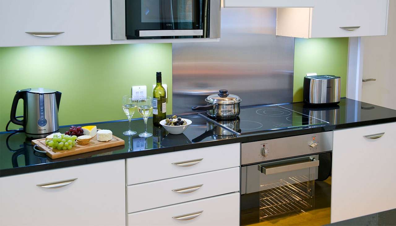 Kitchen counter displays serving items; two wine glasses beside a wine bottle, cheeses with grapes and crackers, olives, kettle, toaster; lidded pot rests on induction hob; white cabinets, green backsplash. Text: PHILIPS, CDA.