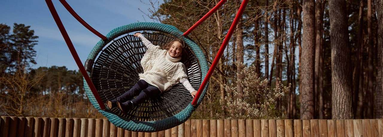 Child on a large circular rope swing reclines and smiles while swinging, held by red ropes, in an outdoor playground bordered by a wooden fence and tall pine trees.