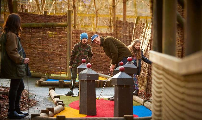 Two adults and two children play miniature golf; one adult putts as the kids watch and smile. Course features colorful turf, wooden obstacles, and woven fences in a woodland setting.