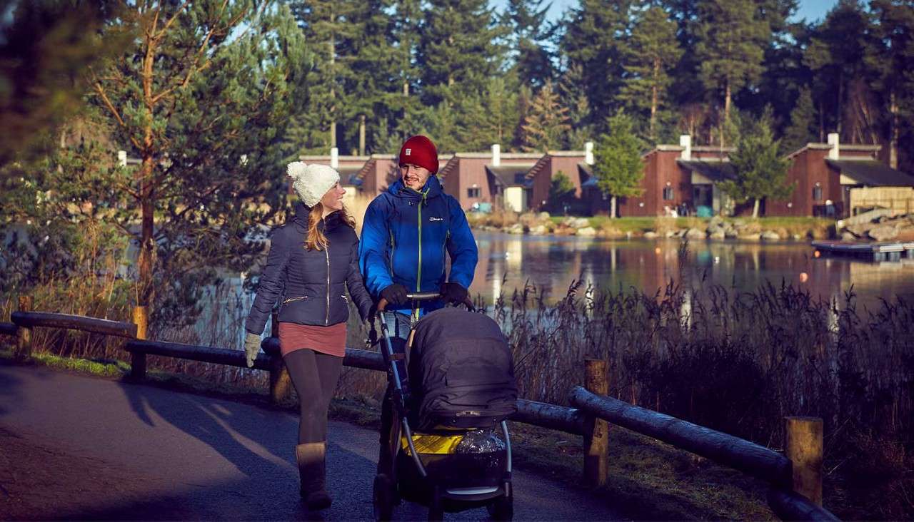 Couple with a stroller walks and chats along a paved lakeside path, wearing winter jackets and hats; wooden cabins and pine trees line the opposite shore under soft afternoon sunlight.
