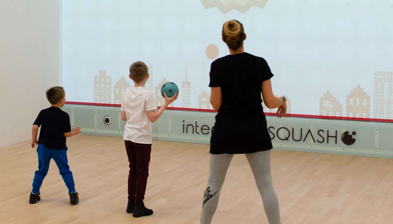 Child holding a teal ball prepares to throw at a digital wall; another child and an adult stand beside in a squash court. Visible text: "inte SQUASH".