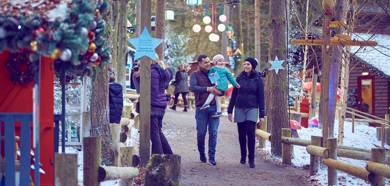 Family with young child—father carrying—walks along a snowy, decorated forest path as others stroll; lanterns and ornaments hang; wooden fences and cabins border the walkway. Text: “Help us keep the magic of Christmas. Please do not walk on the snow.”