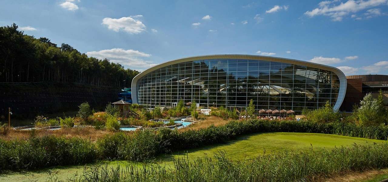 Curved glass-walled building reflecting trees stands beside meandering outdoor water channels. Sun umbrellas line a terrace. Context: landscaped grasses, shrubs, and a forested hillside under a bright, partly cloudy sky.