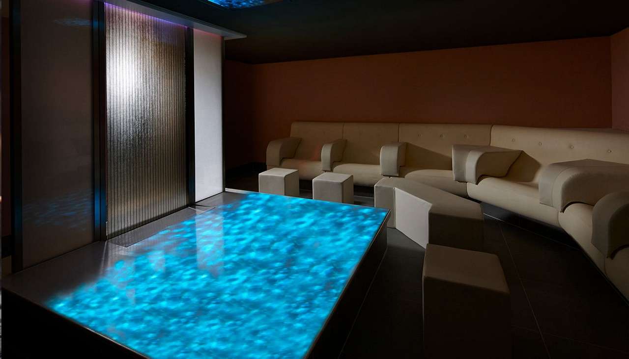 Illuminated glass table glows blue, casting watery reflections. Surrounding it, a modern lounge holds a beige sectional sofa, cube ottomans, and a ribbed glass door under low, dark-ceiling ambient lighting.