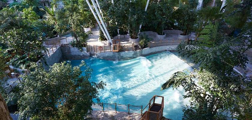 Wave pool generates rolling surf while turquoise water crashes toward a shallow shore; indoor tropical atrium surrounds it with palms, rocky walls, railings, walkways, and tall white support beams overhead.