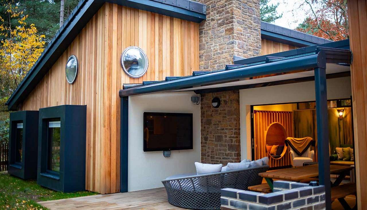 Contemporary wooden house features a covered patio with wall-mounted TV, wicker sofa, and picnic table; circular windows, stone chimney, and warmly lit interior seating appear amid surrounding trees.