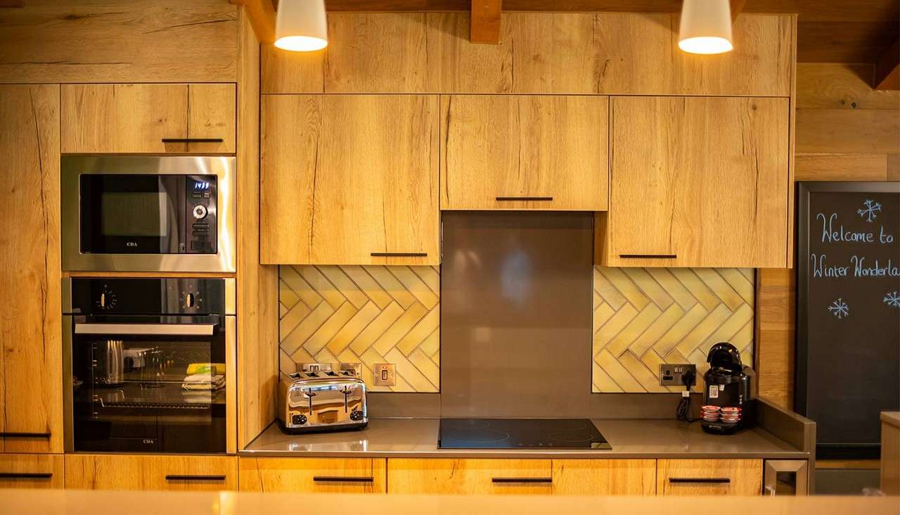 Built-in kitchen appliances line wooden cabinets; toaster and coffee machine on counter; stovetop centered. Warm-lit space with herringbone backsplash and chalkboard reading: "Welcome to Winter Wonderland". Labels: "CDA" on microwave and oven.