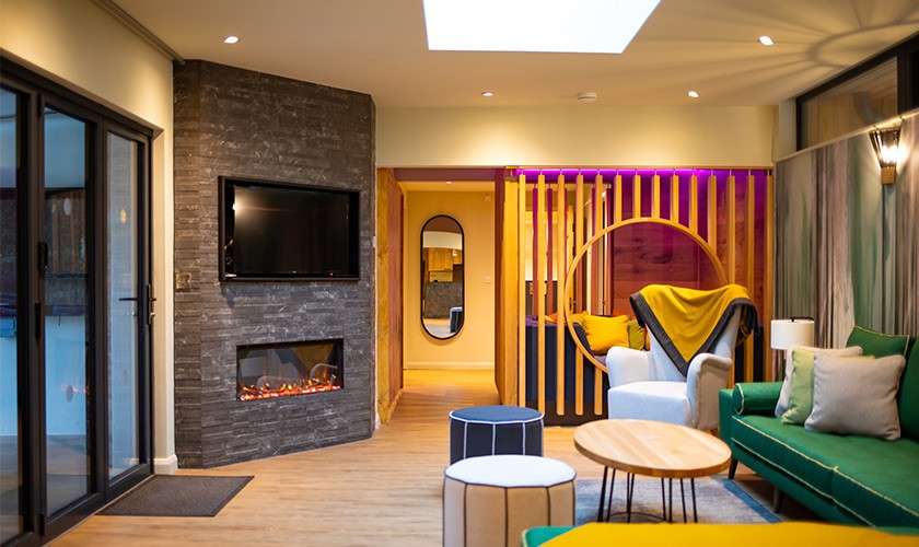 Modern living room features sofas and chairs arranged around tables, illuminated by skylight and recessed lights; TV above electric fireplace; sliding glass doors and partition with circular opening adjoining room.