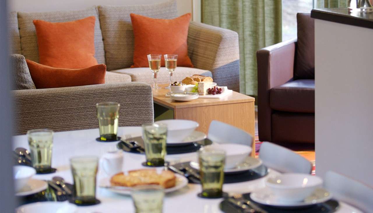 Dining table set with bowls, plates, cutlery, and green-tinted glasses. Nearby sofas with orange cushions flank a side table holding wine, bread, cheese, and grapes in a living room.