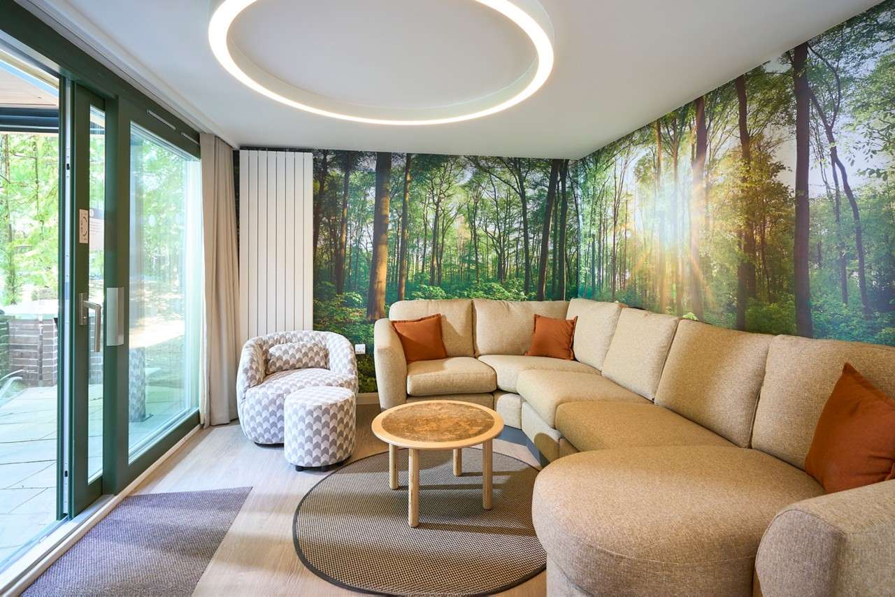 Curved beige sectional sofa anchors the room, encircling a round coffee table, in a bright living space with forest-wall mural, circular ceiling light, patterned chair, and sliding glass doors.