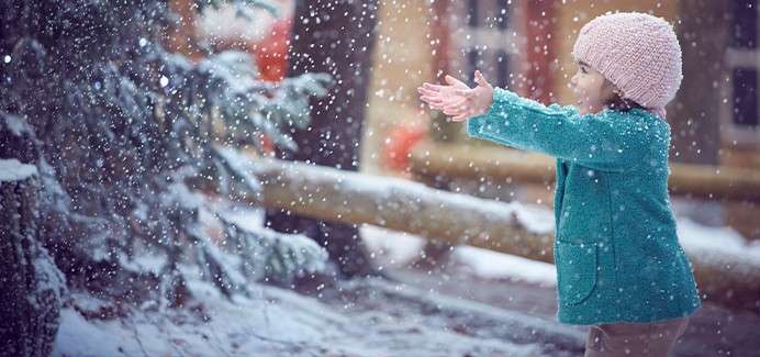 Child reaches out to catch falling snow, standing beside an evergreen and wooden fence, wearing a teal coat and pink knit hat in a snowy outdoor setting.